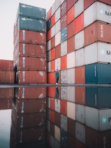 A man walks by towering cargo containers with reflections in the water, capturing the essence of global trade.