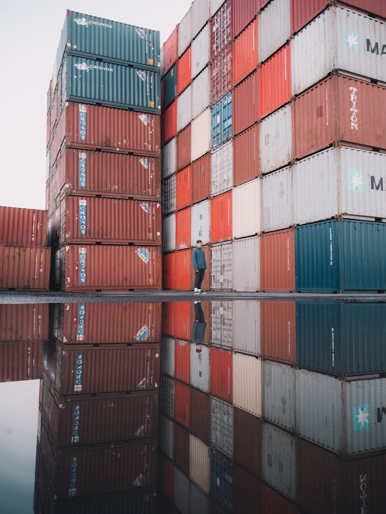 A man walks by towering cargo containers with reflections in the water, capturing the essence of global trade.