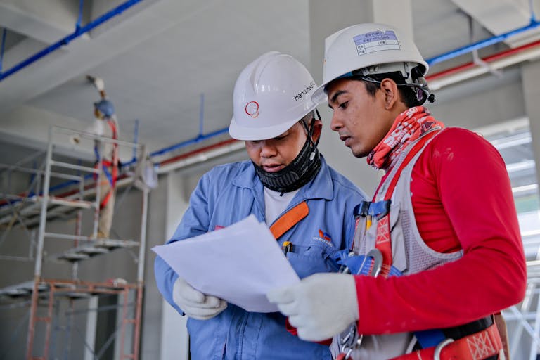 Two engineers in safety helmets reviewing construction plans at a worksite.